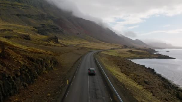 昼間にアイスランドで 雲に覆われた山の斜面を眺めながら川岸近くの山道を走る灰色の車の空中撮影 — ストック動画