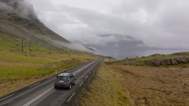 昼間のアイスランドの右側にある川と遠くからの霧に覆われた道路に向かって動く灰色の車の広いショット — ストック動画