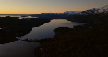 A wide drone flight over the Nahuel Huapi Park and its lush green forest, with a view of the snow-capped mountain range at Bariloche, Argentina during sunrise