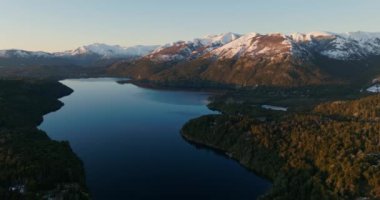 A wide drone flight panning from right to left, over the Nahuel Huapi lake with a view of the sunlit mountain range and lush green forest at Bariloche, Argentina during sunrise