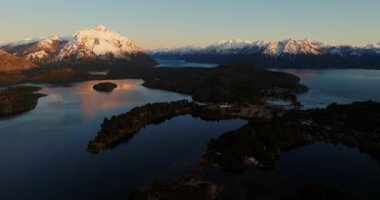 A wide drone flight panning from left to right, over the Nahuel Huapi lake with a view of the sunlit Tronador mountain at Bariloche, Argentina during sunrise