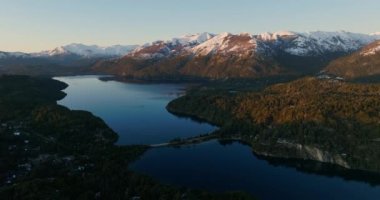 A wide drone flight moving towards, over the Nahuel Huapi lake with a view of the sunlit mountain range and lush green forest at Bariloche, Argentina during sunrise