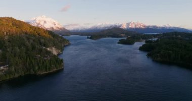 A wide drone flight panning slowly from left to right, over the Nahuel Huapi lake with a view of the sunlit mountain range and lush green forest at Bariloche, Argentina during sunrise