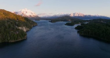 A wide drone flight arcing slowly from left to right, over the Nahuel Huapi lake with a view of the sunlit mountain range and lush green forest at Bariloche, Argentina during sunrise