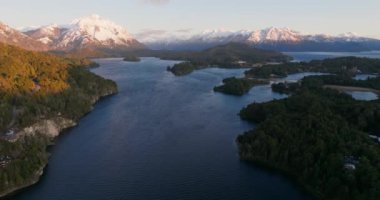 A wide drone flight moving towards, over the Nahuel Huapi lake with a view of the sunlit mountain range and lush green forest at Bariloche, Argentina during sunrise