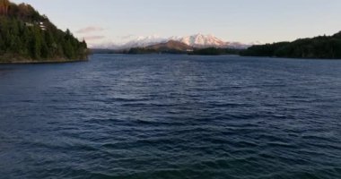 A wide drone flight over the calm waters of the the Nahuel Huapi lake with a view of the sunlit mountain range and lush green forest at Bariloche, Argentina during sunrise