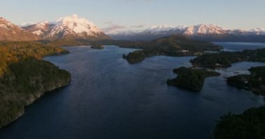 A wide drone flight moving towards, over the Nahuel Huapi lake with a view of the sunlit mountain range and lush green forest at Bariloche, Argentina during sunrise