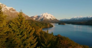 A wide drone flight moving towards, over the lush green forest at Nahuel Huapi park with a view of the sunlit snow-capped mountain range at Bariloche, Argentina during sunrise
