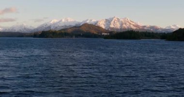 A wide drone flight over the calm waters of the Nahuel Huapi lake with a view of the sunlit mountain range and lush green forest at Bariloche, Argentina during sunrise