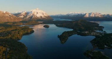 A wide drone flight moving towards, over the Nahuel Huapi lake with a view of the sunlit mountain range and lush green forest at Bariloche, Argentina during sunrise