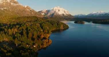 A wide drone flight moving towards, over Nahuel Huapi park and its lush green forest, with a view of the sunlit snow-capped mountain range at Bariloche, Argentina during sunrise
