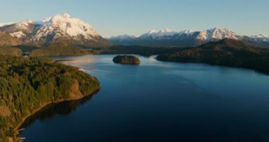A wide drone flight arcing slowly from left to right, over the Nahuel Huapi lake with a view of the sunlit mountain range and lush green forest at Bariloche, Argentina during sunrise
