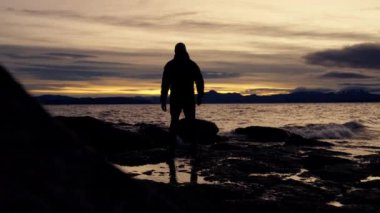 Gün doğarken Arjantin 'in Bariloche şehrinde dalgalar sıçrarken gölde yürüyen ve büyük bir kayanın üzerinde zıplayan bir adamın arka görüntüsünü gösteren geniş bir çekim.