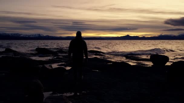 这是一张宽的照片 它展示了一个人在一个岩石巨大的湖滨上行走时的背景色 而日出时 阿根廷巴里洛切的海浪正在飞溅 — 图库视频影像