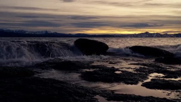 在日出的时候 在阿根廷巴里洛希的一个湖滨 一个从左到右移动着山脉轮廓的平底锅 海浪在岩石上飞溅 — 图库视频影像