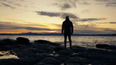 Gün doğarken Arjantin 'in Bariloche şehrinde dalgalar sıçrarken, büyük kayalarla göl kıyısında yürüyen bir adamın arka görüntüsünü gösteren geniş bir çekim.