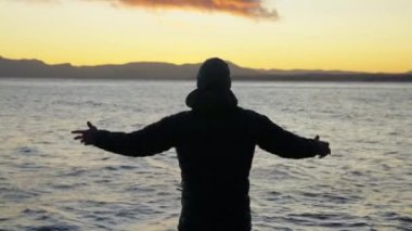Gün doğarken Arjantin 'in Bariloche kentindeki bir göl kıyısında duran ve kollarını kaldıran bir adamın arka görüntüsünü gösteren geniş bir çekim.