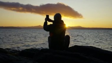 Arjantin 'in Bariloche kentindeki göl kıyısındaki güzel gündoğumunun panoramik görüntüsünü yakalamak için cep telefonunu kullanan bir adamın arka görüntüsünü gösteren geniş bir çekim.