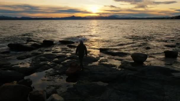 在阿根廷巴里洛切 一架无人驾驶飞机拍摄到一名男子沿着岩石海岸向湖面走去 朝日出方向望去 — 图库视频影像