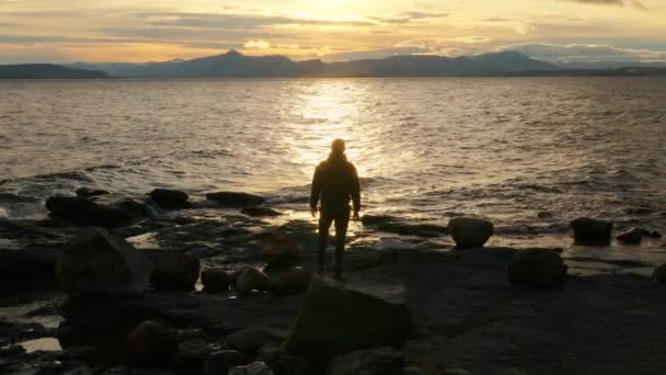 アルゼンチンのバリローチェで日の出の間に湖の景色を眺めながら腕を上げている男のシルエットの広いショット — ストック動画