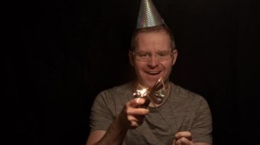 A medium shot of a man with a silver party hat, bow tie, and safety glasses, smiling and doing hand movements while holding sparklers, against a dark background