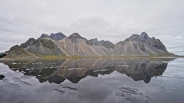 Vestrahorn sıradağlarının geniş bir görüntüsü ve İzlanda 'da bulutlu bir günde dağın yansımasını gösteren bir adam suyun üzerinde yürüyor.