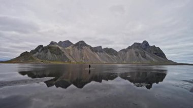 Vestrahorn dağından uzaklaşan bir adamın geniş bir görüntüsü ve sakin sular İzlanda 'da bulutlu bir günde dağın yansımasını gösteriyor.