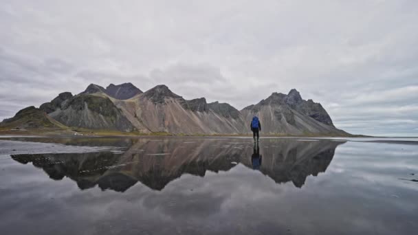 アイスランドの曇りの日に山の反射を示すヴェストラホーン山と水に向かって歩く男の広いショット — ストック動画
