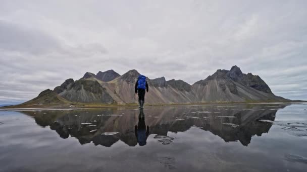 アイスランドの曇りの日に山の反射を示す水の上を歩いて フレームの左側から来る男とヴェストラホーン山脈の広いショット — ストック動画
