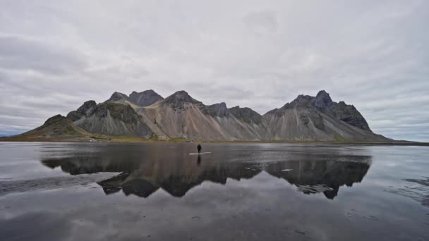 アイスランドの曇りの日に山の反射を示すヴェストラホーン山とまだ水から歩いて男の広いショット — ストック動画