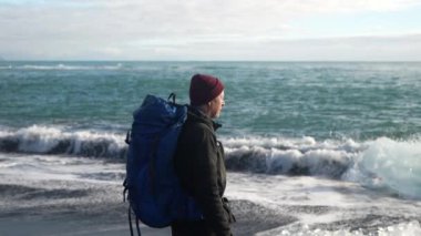 Slow motion footage of a man carrying a backpack standing near an iceberg fragment on the shore of Diamond Beach on a cloudy day