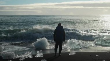Back view slow motion footage captured at sunset of a man facing the approaching waves at Diamond Beach with nearby ice chunks