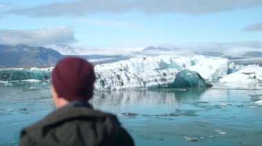 İzlanda 'da gündüz vakti Jokulsarlon Buzul Gölü' ne odaklanan bir adamın bulanık sırtından sağa sola hareket eden bir çekim.