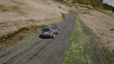 A daytime wide shot of a gray car traveling on a rocky road with a view of the mountain in Iceland