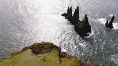 A daytime aerial shot of a man standing on a cliff overlooking the Reynisdrangar sea stacks