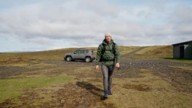 A video showing a man walking on a field towards the camera with a gray car and house in the background during daytime in Iceland
