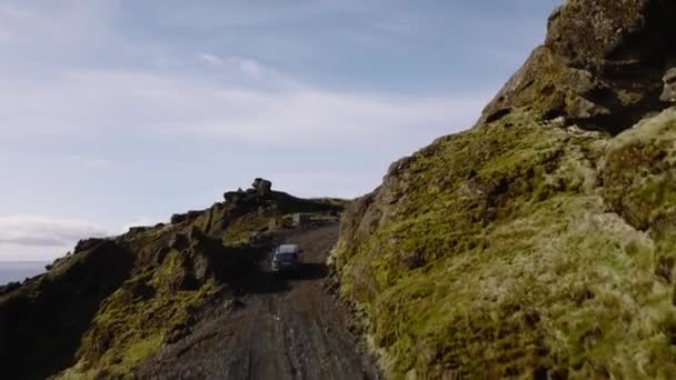 灰色の車の昼間の空中ショットは アイスランドの風景と海を眺めながら 草や岩の多い山の横の道路を走行します — ストック動画