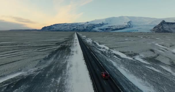 昼間のアイスランドの雪に覆われた山脈の景色を望む道路上で冒険する赤い車の広い空中ショット — ストック動画