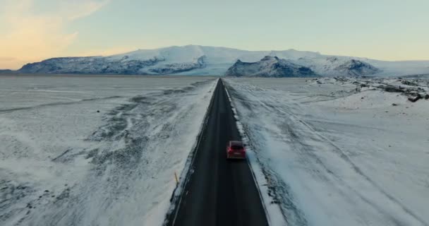 昼間にアイスランドで雪に覆われた道端や山の風景と一緒に道路を走行する1台の赤い車の空中追跡ショット — ストック動画
