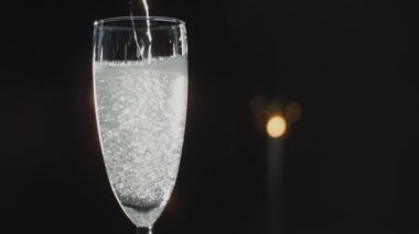 Bir flüt bardağının yakın plan çekimi, şampanya yavaş çekimde, parlak ışık alan koyu bir arka plana dökülüyor.