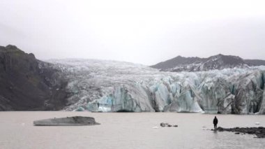 Gündüz vakti, kayalık bir göl kıyısında, İzlanda, Skaftafell 'de gündüz vakti Svinafelljokull buzulu ile sisli manzaraya bakan bir adamın görüntüsü.