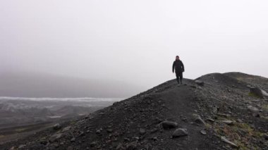 Skaftafell, İzlanda 'da gündüz vakti Svinafellsjokull buzulunun sisli manzarasıyla kayalık bir arazide yürüyen bir adamın geniş açılı görüntüsü.