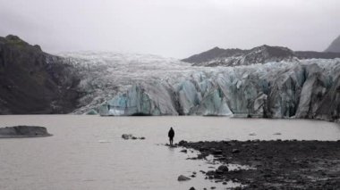 Gündüz vakti kayalık bir göl kıyısında duran bir adamın Svinafelljokull buzulu ve İzlanda 'nın Skaftafell kentinde gündüz vakti bir dağdan oluşan puslu manzaraya bakışı.