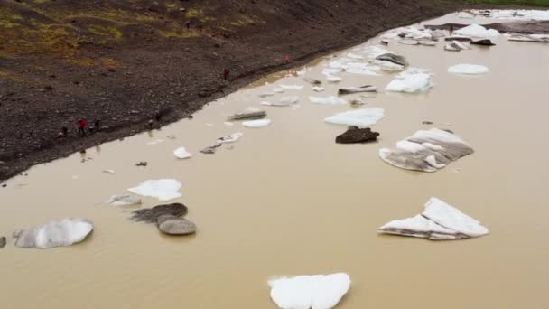 アイスランドのスカッタフェルにある茶色の川に浮かんでいる スヴィネフェルスジョクル氷河の氷の塊を見て 岩の多い湖畔の人々の上にドローン — ストック動画