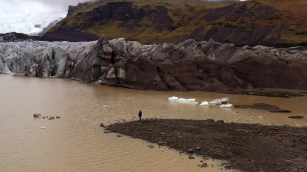 アイスランドのスカフタフェルにあるSvinafellsjokull氷河を見て 岩の多い湖畔の端に立っている男に向かって移動する空中ドローンショット — ストック動画