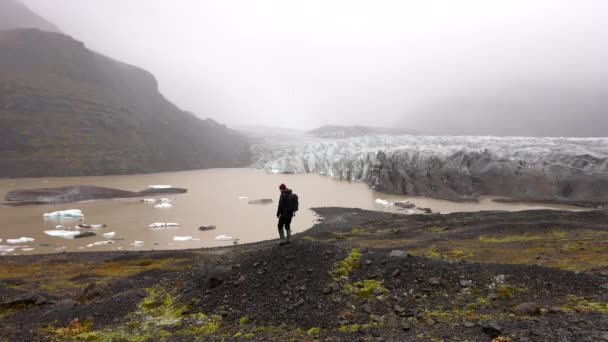 アイスランドのSkaftafellで昼間に霧の多い風景とSvinafellsjokull氷河に対して岩の多い湖畔を歩く男の昼間のワイドショット — ストック動画