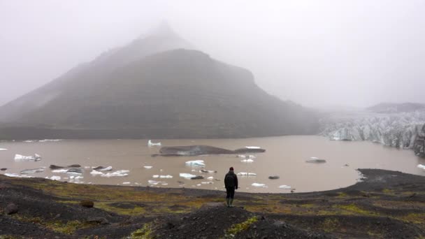 岩だらけの湖畔に立っている男の昼間のワイドショット アイスランドのSkaftafellで昼の間にSvinafellsjokull氷河と山からなる霧の風景を見て — ストック動画
