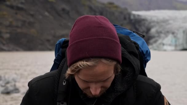 アイスランドのSkaftafellで昼間の背景にSvinafellsjokull氷河と山で カメラをまっすぐに見て 彼の頭を持ち上げる男のクローズアップショット — ストック動画