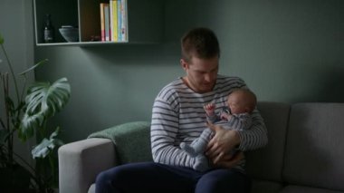 Bir adam kanepeye oturur, ninni söyler ve nazikçe kucağında bir bebek sallar.