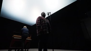 Bir şarkıcıyla piyanistin, karanlık bir arka planı olan bir stüdyonun parlak ışıkları altında müzik yaptıkları düşük açılı çekimler.